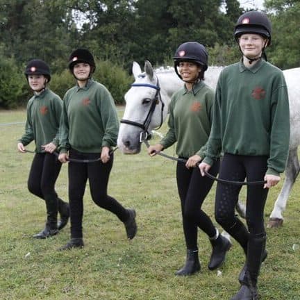 Four Horse Rangers in green jumpers and riding hats walking a dappled grey horse across the field.