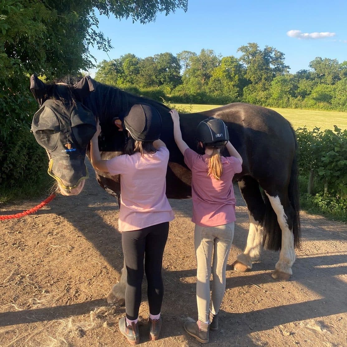 Two Horse Rangers in riding hats smoothing a rug over a black and white horse on a summer evening.